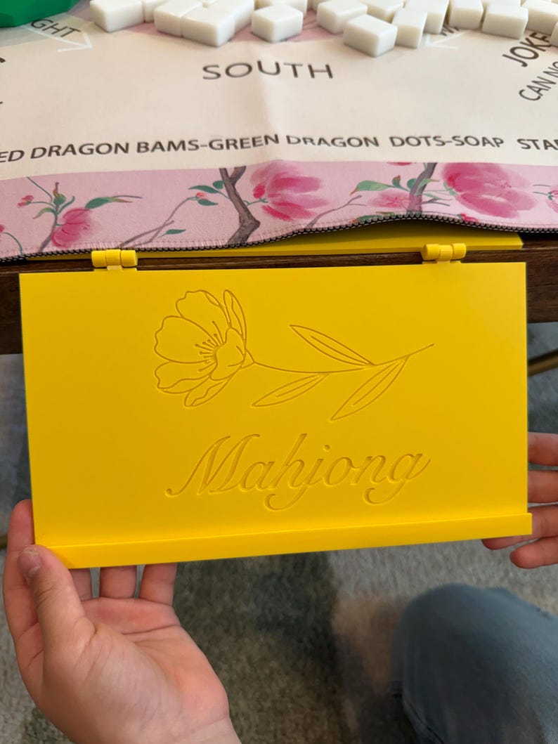 May include: A bright yellow rectangular Mahjong tile rack featuring a hinged design. The front surface is engraved with a delicate line-art illustration of a single flower with leaves and the word Mahjong written in a cursive script font.