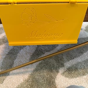 May include: A bright yellow rectangular box with a hinged lid. The front panel features an engraved line art illustration of a single flower with leaves and the word Mahjong written in a cursive script font centered below the floral design.