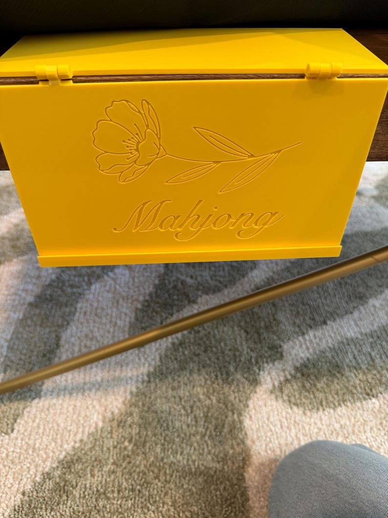 May include: A bright yellow rectangular box with a hinged lid. The front panel features an engraved line art illustration of a single flower with leaves and the word Mahjong written in a cursive script font centered below the floral design.