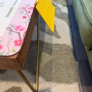 May include: A rectangular table with thin gold metal legs and a light pink surface featuring a floral pattern and text that reads DRAGONS CRAKS-RED DRAGON BAMS-GREEN DRAGON. A bright yellow plastic card holder with an embossed floral design is attached.