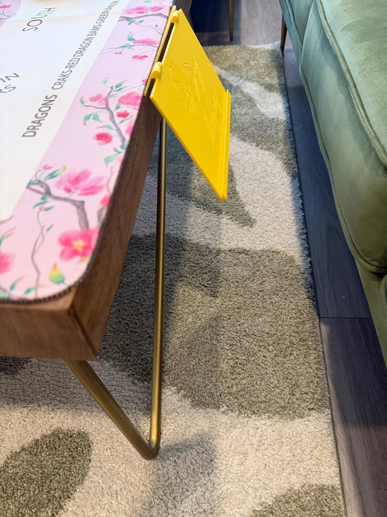 May include: A rectangular table with thin gold metal legs and a light pink surface featuring a floral pattern and text that reads DRAGONS CRAKS-RED DRAGON BAMS-GREEN DRAGON. A bright yellow plastic card holder with an embossed floral design is attached.