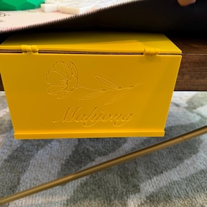 May include: A bright yellow plastic Mahjong tile rack with a hinged design that hooks over the edge of a table. The front panel features an engraved line art illustration of a single flower and the word Mahjong in a cursive script font.