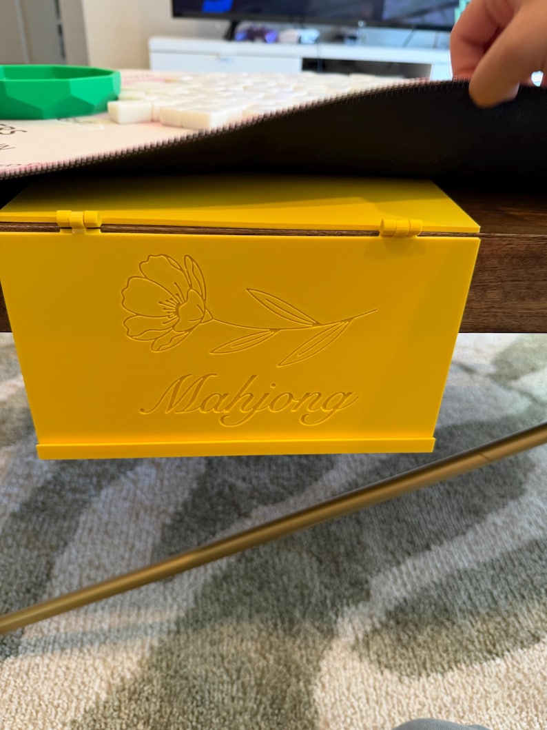May include: A bright yellow plastic Mahjong tile rack with a hinged design that hooks over the edge of a table. The front panel features an engraved line art illustration of a single flower and the word Mahjong in a cursive script font.