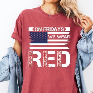 May include: Red, white, and blue distressed American flag graphic on a maroon t-shirt. The text "On Fridays We Wear Red" is written below the flag. The words "Remember", "Everyone", "Deployed" are written vertically in white on a red background.