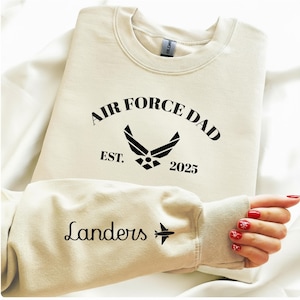 May include: Cream-colored sweatshirt with the text "AIR FORCE DAD" above an Air Force emblem and the year "2025". The left sleeve has the name "Landers" and a small airplane graphic. The sweatshirt is made of a soft material.