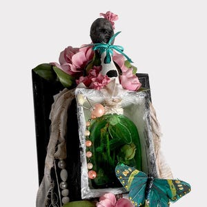 May include: A framed shadow box containing a green glass bottle, pink flowers, a butterfly, and a pearl necklace. The bottle is filled with a green liquid and has a white stopper. The butterfly is blue and green with black spots. The flowers are pink and white.