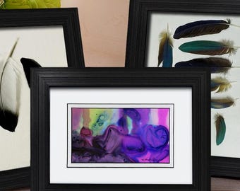 May include: Three black rectangular picture frames arranged together. The left frame displays a single white feather with a black tip. The right frame shows a collection of colourful feathers. The centre frame contains a small, vibrant abstract print.