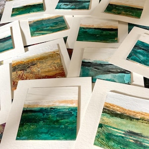 以下が含まれることがあります： 白い紙の台紙に載せられた小さな正方形の絵画のコレクション。層状のテクスチャを持つ抽象的な風景が描かれています。作品には、深いティール、フォレストグリーン、オーカー、白の絵の具の水平な帯があり、さまざまな地平線が表現されています。