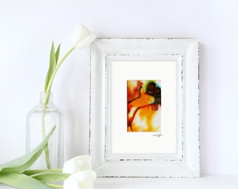 May include: A white distressed wooden frame containing an abstract painting with vibrant orange, yellow, and teal ink splashes. The artwork is matted in white and features a signature in the bottom right corner, displayed next to a clear glass vase with white tulips.