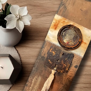 May include: A decorative arrangement featuring a gray geometric candle, a small white vase with white flowers, and a rustic wooden piece with metal accents and a tassel. The items are arranged on a wooden surface.