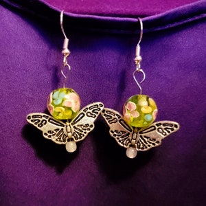 Puede incluir: Un par de pendientes de mariposa plateados con cuentas de vidrio florales verdes y rosas. Las mariposas tienen alas detalladas y las cuentas tienen un patrón moteado.