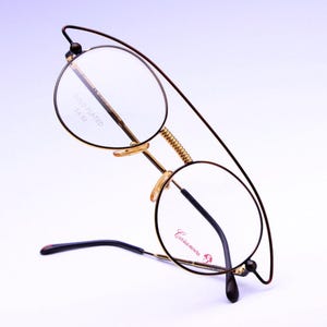 May include: Gold-plated round eyeglasses with black frames and a spring hinge. The glasses have a Casanova logo on the right lens and the text "Gold Plated 24 Kt" on the left lens.