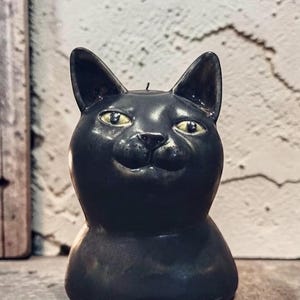 May include: A black cat-shaped candle with a smooth, glossy finish. The candle features pointed ears, a small nose, and two yellow eyes with black pupils. A single black wick is visible at the top of the cat's head, centered between the ears.