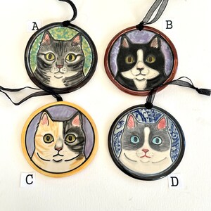 May include: Four circular ceramic cat ornaments labelled A, B, C, and D. Ornament A features a grey tabby cat, B a black and white cat, C a calico cat, and D a grey and white cat. Each ornament has a black ribbon for hanging and unique patterns.