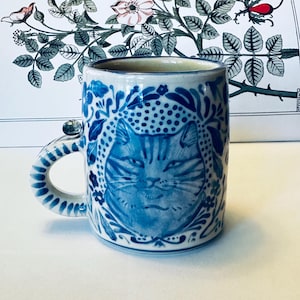 May include: A ceramic mug featuring a hand-painted blue and white cat portrait surrounded by intricate floral and leaf patterns. The handle is decorated with blue stripes and a small, spherical accent piece attached to the top rim area.