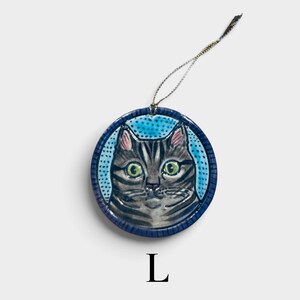 May include: A round ceramic ornament featuring a hand-painted grey tabby cat with large green eyes against a light blue dotted background. The circular piece has a dark blue border and includes a thin metallic gold hanging string at the top.