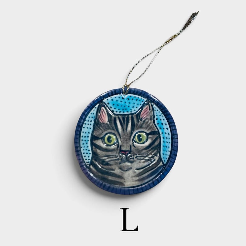 May include: A round ceramic ornament featuring a hand-painted grey tabby cat with large green eyes against a light blue dotted background. The circular piece has a dark blue border and includes a thin metallic gold hanging string at the top.