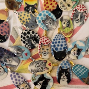 May include: A collection of oval-shaped pendants featuring hand-painted cat faces. The pendants showcase various cat breeds and patterns, with backgrounds of polka dots, hearts, and solid colors. Each pendant has a silver bail for hanging.