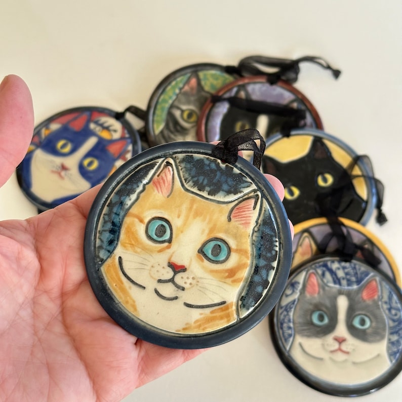 May include: A collection of round ceramic ornaments featuring hand-painted cat faces in various colours and patterns. Each circular piece is framed by a dark blue border and includes a black ribbon loop at the top for hanging purposes.