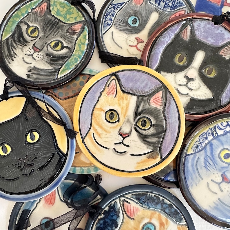 May include: A collection of round ceramic ornaments featuring hand-painted cat portraits. Each piece displays a unique feline face with detailed expressions, vibrant colours, and black ribbon loops for hanging. The set includes various cat breeds.