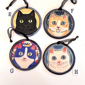 May include: Four circular ceramic ornaments labelled E, F, G, and H, each featuring a cat portrait. E shows a black cat on yellow, F an orange tabby on blue, G a blue and white cat on a patterned background, and H a white cat on blue. Each has a black ribbon.