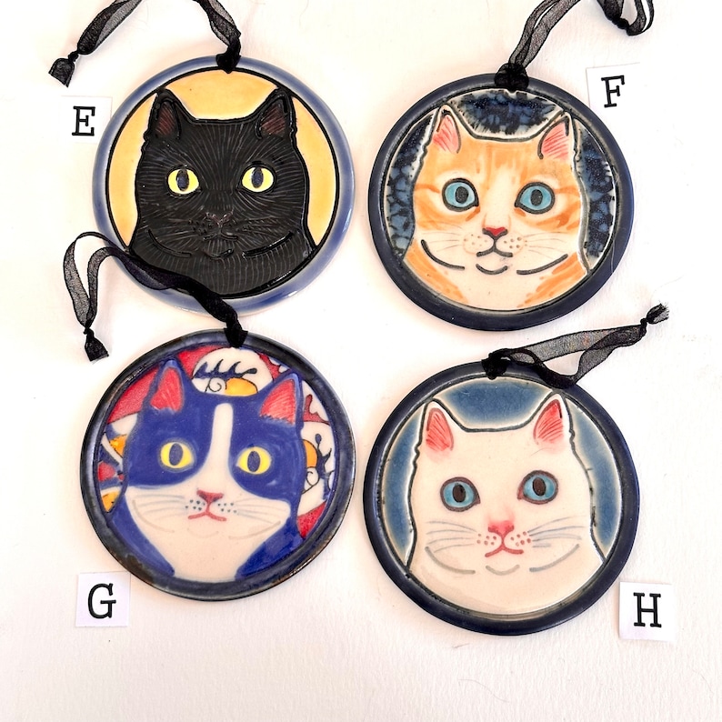May include: Four circular ceramic ornaments labelled E, F, G, and H, each featuring a cat portrait. E shows a black cat on yellow, F an orange tabby on blue, G a blue and white cat on a patterned background, and H a white cat on blue. Each has a black ribbon.