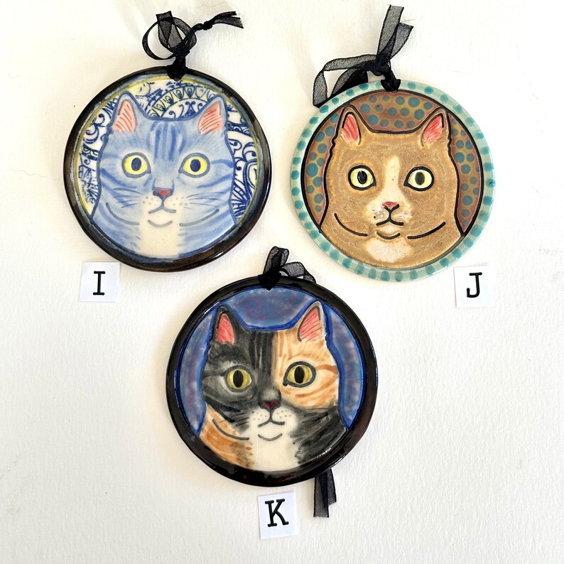 May include: Three circular ceramic ornaments featuring cat portraits labelled I, J, and K. Ornament I shows a blue tabby cat, J shows a tan cat with a blue dotted background, and K shows a calico cat. Each has a black ribbon for hanging.