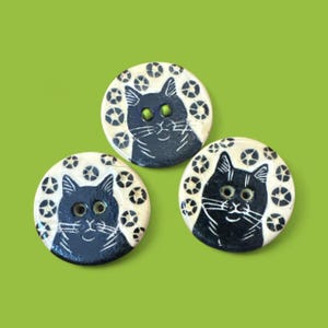 May include: Three round, handmade buttons with a black cat design on a cream background. The cats have detailed white whiskers and are surrounded by a pattern of small, stylized shapes. The cat's eyes are green. The background is a solid, bright green.
