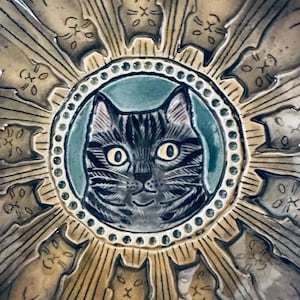 May include: A round ceramic bowl featuring a central circular medallion with a detailed gray tabby cat face on a teal background. The rim is decorated with a ring of small dots, surrounded by a radial pattern of twelve stylized cat figures.