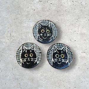 May include: Three round, black ceramic buttons with white cat faces. Each button features a smiling cat face with large eyes and a grid-like pattern around the edge. The buttons are arranged on a gray surface.