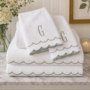 May include: A stack of white towels featuring a sage green scalloped edge embroidery along the bottom hem. The top two towels are folded to display a sage green embroidered letter G monogram centered above the scalloped decorative border detail.