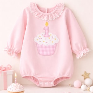 May include: Pink baby romper with a cupcake applique featuring a number one candle. The long-sleeved romper has a ruffled collar and cuffs. The cupcake has a gingham pattern. A birthday theme.