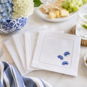 Puede incluir: Servilletas cuadradas de lino blanco con dise&ntilde;o de cangrejo azul. Las servilletas tienen un borde cosido. La imagen tambi&eacute;n incluye un jarr&oacute;n floral azul y blanco, un vaso de agua y un plato de aceitunas. Servilletas sobre una superficie blanca.