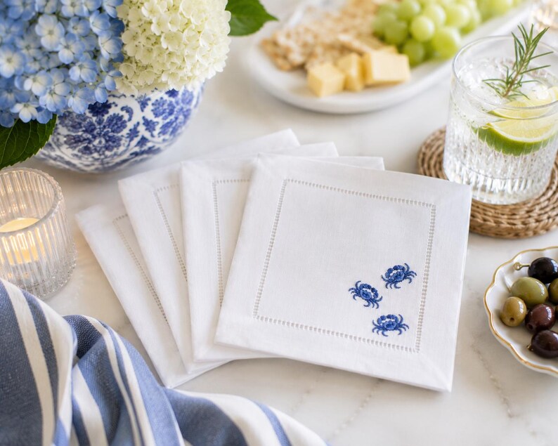 Puede incluir: Servilletas cuadradas de lino blanco con dise&ntilde;o de cangrejo azul. Las servilletas tienen un borde cosido. La imagen tambi&eacute;n incluye un jarr&oacute;n floral azul y blanco, un vaso de agua y un plato de aceitunas. Servilletas sobre una superficie blanca.