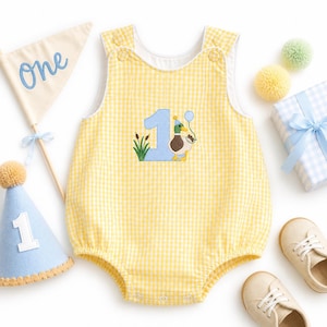 May include: A yellow and white gingham romper featuring an embroidered light blue number one, a mallard duck wearing a party hat, and cattails. The romper is surrounded by a pennant flag with the word One, a blue party hat, and beige shoes.