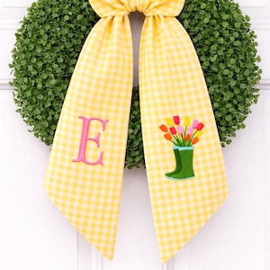 May include: A decorative wreath with a green, leafy base. A yellow gingham ribbon is attached, featuring a pink embroidered "E" and a colorful tulip design with green rain boots. The wreath is likely for spring or Easter.