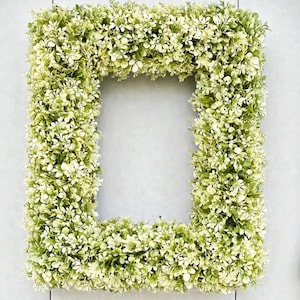 May include: A rectangular frame covered in small, light green and white faux leaves. The frame is a square shape with a large, open center. The leaves are densely packed, creating a textured surface.