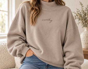 Könnte beinhalten: Ein beigefarbenes Sweatshirt mit Stehkragen und dem Wort worthy, das in dunkelbrauner Schreibschrift mittig auf der Brust eingestickt ist. Das Kleidungsstück hat lange Ärmel mit gerippten Bündchen und eine lockere Passform, kombiniert mit einer blauen Jeans.