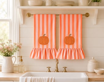 May include: Two kitchen towels hanging on a brass rod, featuring vertical stripes in alternating shades of coral orange and light pink. Each towel has a ruffled bottom hem and a centered embroidered orange pumpkin design with a brown stem.