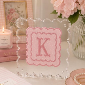 May include: A clear acrylic frame with a scalloped edge holds a pink fabric square featuring a white polka dot pattern and a scalloped border. Centered on the fabric is a large, embroidered letter K in a matching shade of pink thread.