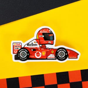 Könnte beinhalten: Eine Cartoon-Illustration eines roten und weißen Formel-1-Rennwagens mit einem Fahrer, der einen roten Helm mit dem Wort "Marlboro" trägt. Das Auto trägt die Aufschriften "Marlboro", "Shell", "AMD" und "Vodafone".