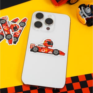 Puede incluir: Un iPhone blanco con una pegatina de coche de carreras rojo, blanco y azul en la parte posterior. La pegatina presenta un piloto de carreras de dibujos animados con un casco rojo con la palabra "Marlboro" en el lateral del coche.