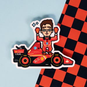 Può includere: Un'auto da corsa rossa e nera con un pilota di cartoni animati che alza le braccia in segno di vittoria. L'auto ha la scritta "Ray Ban" sul lato e "IGK" sul retro.