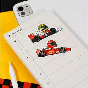 Puede incluir: Dos pegatinas de coches de carreras de dibujos animados en un planificador semanal blanco. La pegatina superior es un coche de carreras blanco y rojo con un logotipo de Marlboro y el n&uacute;mero 2. La pegatina inferior es un coche de carreras rojo con un logotipo de Marlboro y el n&uacute;mero 5.