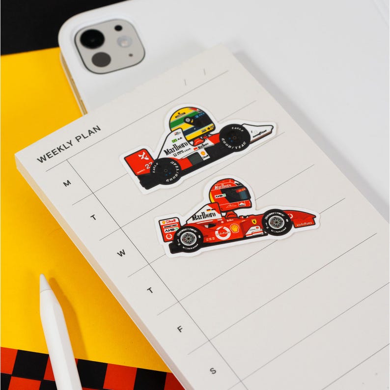 Puede incluir: Dos pegatinas de coches de carreras de dibujos animados en un planificador semanal blanco. La pegatina superior es un coche de carreras blanco y rojo con un logotipo de Marlboro y el n&uacute;mero 2. La pegatina inferior es un coche de carreras rojo con un logotipo de Marlboro y el n&uacute;mero 5.
