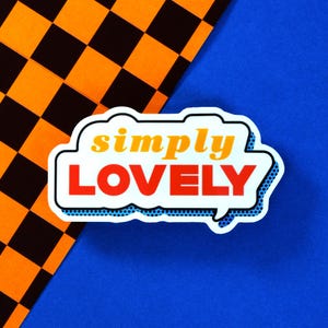 Könnte beinhalten: Eine weiße Sprechblase mit einem blauen gepunkteten Rand und dem Text "simply LOVELY" in orangefarbenen und roten Buchstaben. Die Sprechblase befindet sich auf einem blauen Hintergrund mit einem schwarz-orangenen Karomuster im Hintergrund.