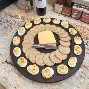 May include: A round wooden serving tray with a variety of snacks, including deviled eggs, crackers, and a block of cheese. The tray is decorated with a knife and a small amount of red pepper flakes.