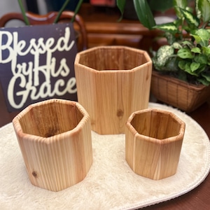 Può includere: Tre vasi da fiori ottagonali in legno di diverse dimensioni. I vasi sono in legno naturale e hanno un colore marrone chiaro. I vasi sono posizionati su un tappeto ovale bianco.