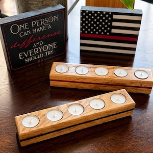 May include: Two rectangular wooden candle holders, each with five circular cutouts containing white tea light candles. One holder features a thin red horizontal stripe, and the other has a thin black stripe. Behind them are two black signs.
