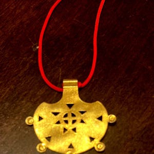 May include: Gold-colored pendant with a geometric design and a red cord. The pendant has a central starburst pattern and triangular cutouts. The cord is a vibrant red, creating a striking contrast with the pendant's color.